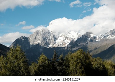 The view of snow-capped Mountain with evergreen trees in foreground. The panoramic view of mountains, blue sky in sunlight and white steam clouds.