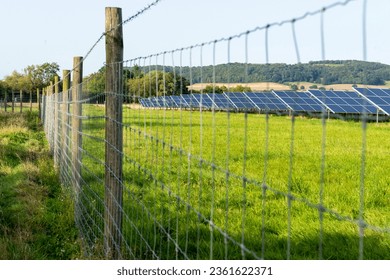 View of solar panels,beyond barbed wire,absorbing sunlight,amongst green summertime grass,in the English countryside.