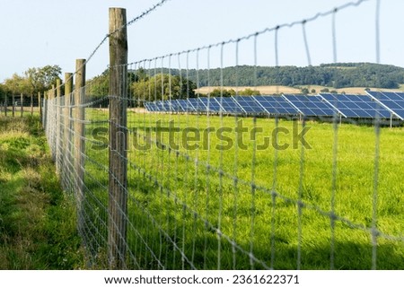 View of solar panels,beyond barbed wire,absorbing sunlight,amongst green summertime grass,in the English countryside.