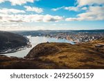 view of St. John’s harbor with cityscape and ocean under a blue sky with clouds. Rolling hills in the foreground add depth, showcasing the natural beauty and urban charm of St. John’s, Newfoundland. 