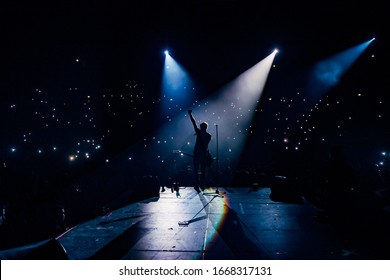 View from the stage stadium. Vocalist of a popular pop band on the background of the flashlights  of phones during a concert. Fans switch on lights on smart phones on tribunes. Crowd waving cellphones - Shutterstock ID 1668317131