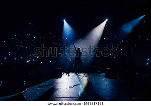 View from the stage stadium. Vocalist of a\
popular pop band on the background of the flashlights  of phones\
during a concert. Fans switch on lights on smart phones on\
tribunes. Crowd waving\
cellphones