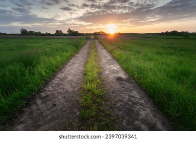 View of a straight and empty dirt road through green meadows with sunset, May evening - Powered by Shutterstock