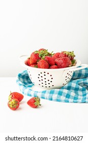View of strawberries in a white colander on a white table with a blue kitchen cloth, white background with three strawberries, vertical