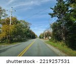 The view of a street leading out of Lockeport, Nova Scotia on a sunny autumn day. Trees and houses line either side of the street with the leaves changing color on many of the trees.