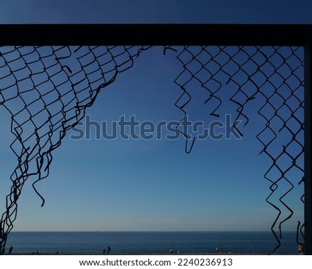 View of the summer beach through a torn metal mesh net fence. black sea coast from behind metal wire fence.