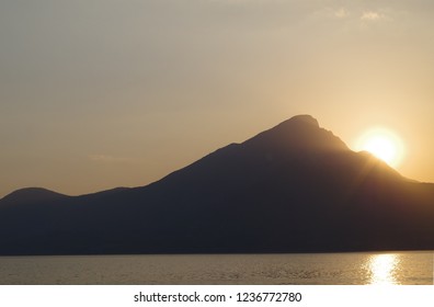 A view of a sun setting behind a mountain forming a silhouette on a lake