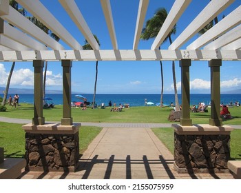 View of a sunny beach from a white wooden shelter. Symmetrical shadows opposite to the roof. Deep blue sea, clear blue sky, palm trees, green lawn, stone pillars, people sunbathing and relaxing. 