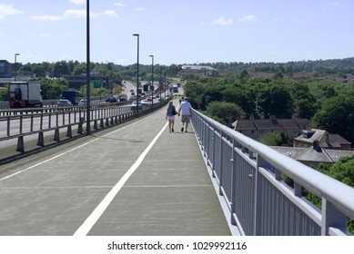 View From A Suspension Bridge, A Couple Walking Away From Camera In Far Mid Ground.      