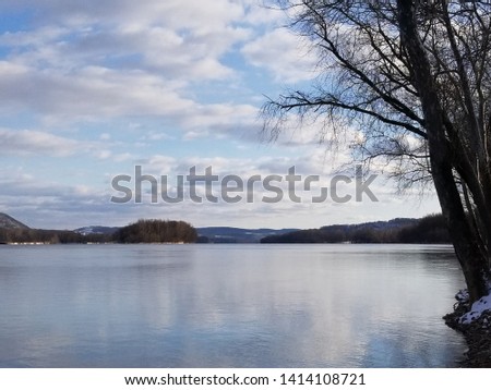 A view of the Susquehanna River reflecting the cloudy sky, with trees to the right hand side amidst a snow covered landscape with Mountain ridges in the background