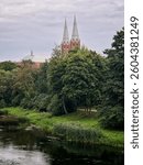 View of Sventoji river and Church of Apostle Evangelist St. Matthew in Anyksciai, Lithuania. A serene river bordered by lush greenery flows through a landscape. 