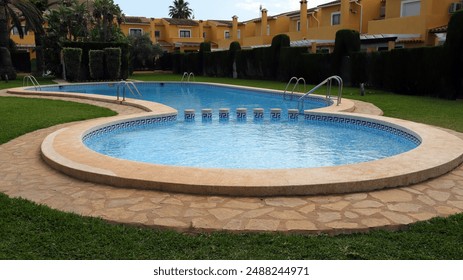 View of swimming pool with unique curved shape, surrounded by lush green grass and stone pavers. Pool is set against backdrop of Mediterranean-style villas with terracotta roofs and lush greenery. - Powered by Shutterstock