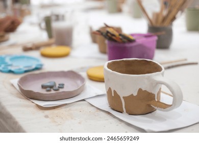 View of the table in a pottery workshop during a master class. On the table there is a mug and a plate made of clay by one of the beginning students. The products are decorated with colorful flowers.