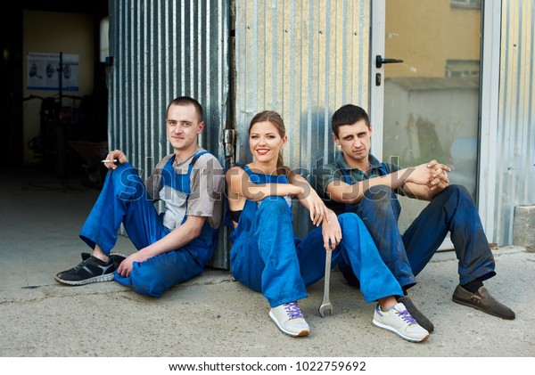 View of a team of\
mechanics sitting near the repair garage. Girl holds a wrench in\
her hand, she sitting between two guys in work clothes and they\
looking at the camera.