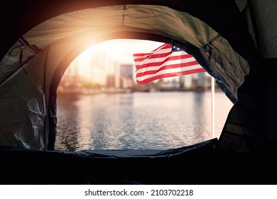 View From A Tent Of A Homeless On A Waving Flag Of USA And Down Town Out Of Focus In The Background At Sunrise. Social Issue, Inadequate Affordable Housing And Income Supply Concept. Sun Flare.