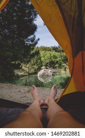 view from a tent on sunset at beach, human legs lying in tourist tent with view of the lake in the forest.
