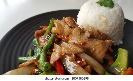 View Of Thai Style Stir Fry Pork Fillet And Mixed Seasonal Vegetables (long Bean, Snow Peas, Capsicum, Chinese Broccoli) With White Jasmine Rice Serve On Black Ceramic Plate. 