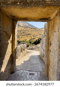 View through an ancient stone passageway at the Mycenae archaeological site in Greece, showing rugged hills and golden summer landscape under bright midday sunlight.
