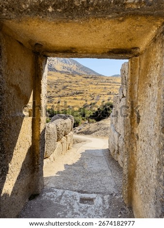 View through an ancient stone passageway at the Mycenae archaeological site in Greece, showing rugged hills and golden summer landscape under bright midday sunlight.