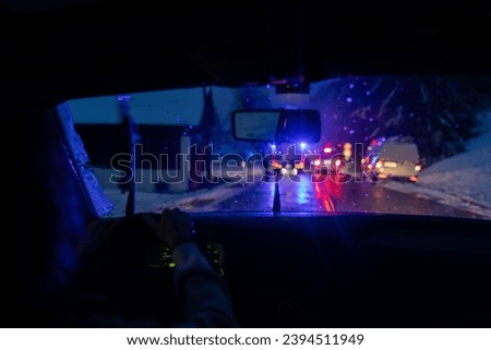 View Through Car Windshield Reveals a Traffic Accident Scene with Flashing Blue Police and Red First Aid Ambulance Lights, Capturing the Urgency and Challenges of Slippery Winter Roads