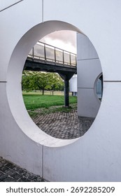 View Through a Circular Architectural Opening onto a Pedestrian Bridge, Green Park, and Modern Building in an Urban Setting
