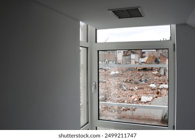 View through a closed window of a construction site with an excavator and piles of construction debris and bricks.