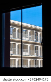 View through a dark interior window framing the sunlit facade of a modern apartment building with repeating balconies and railings against a clear blue sky, suggesting calm urban minimalism and geometry.