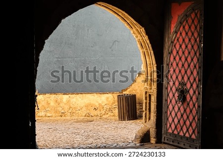 View through a dark, weathered stone archway into a cobblestone courtyard with a textured wall and a striking red diamond-patterned door