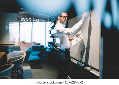 View Through Glass Of Busy Male In Formal Clothes And Glasses Writing Information On Whiteboard Creating Project In Modern Conference Hall