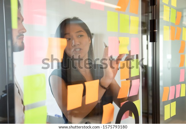 View through glass partition asian female\
worker working strategizing with caucasian male colleague, attached\
colourful post-it notes on glass wall where written ideas actions\
plan for aim achievement
