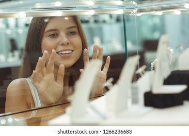 View through glass of pretty young female customer looking at new accessories in showcase in jewelry store. Attractive lady keeping with hands glass and admiring luxurious jewellery. - Powered by Shutterstock