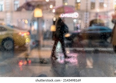 View Through Glass Window With Rain Drops On Blurred Reflection Silhouettes Of The People In Walking On A Rain Under Umbrellas And Bokeh City Lights, Night Street Scene.