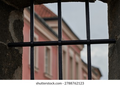 View through iron window bars in old stone wall, blurred pastel building and cloudy sky in background, conveying confinement, security, and historic architecture.