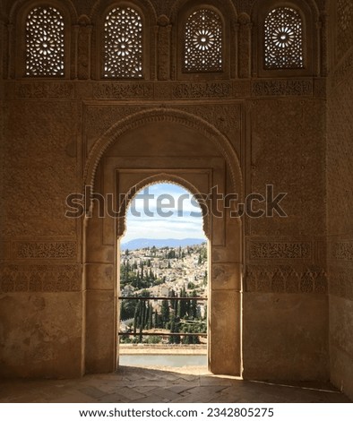 View through Islamic or Moorish style archway to town in Alhambra Spain. Ornate stonework frames the archway with view of green trees, Spanish houses, mountains and blue sky in the distance. 