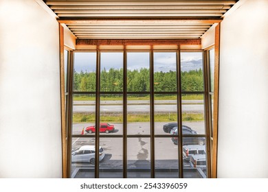 View through large window showing parking lot with  cars, road and forest under clear sky from modern building. Sweden.