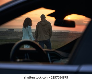 View through open side and front car window of couple in love. Back view of romantic pair looking at evening sky in orange and red colors while sunset out of city.