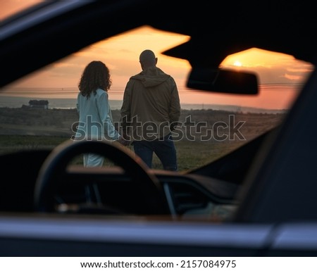 View through open side and front car window of couple in love. Back view of romantic pair looking at evening sky in orange and red colors while sunset out of city.