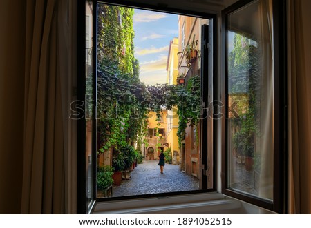 View through an open window as a young woman walks through an ivy covered colorful alley in the Trastevere district of Rome, Italy