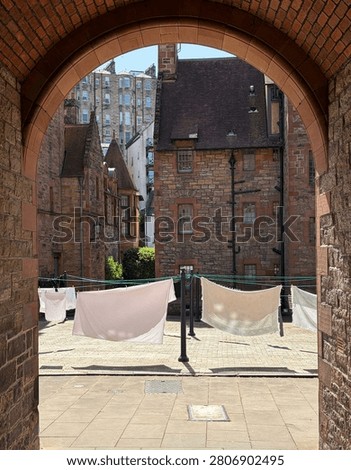 View through a red sandstone archway into a historic cobbled courtyard in Dean Village, Edinburgh, Scotland. White laundry hangs on washing lines strung between Victorian cast iron lamp posts. Traditional red sandstone and ashlar stone buildings with pitched roofs and period windows surround the courtyard. Blue sky visible above the rooftops. No people visible.