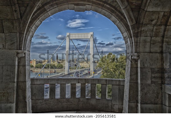 A view through\
round windows of rock balcony to the  dramatic tall white pillars\
of windy elisabeth bridge in Budapest on the Danube River under a\
deep dark blue cloudy sky
