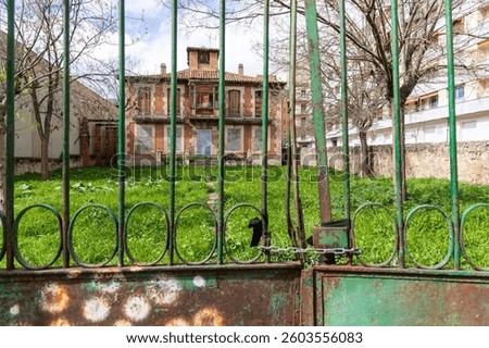 View Through Rusty Locked Iron Gate of Abandoned Red Brick House with Overgrown Garden in Salamanca, Spain Surrounded by Urban Buildings