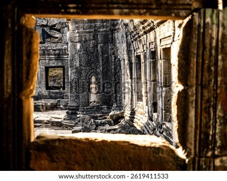 A view through a stone frame showcasing ancient temple ruins with intricate carvings and a statue. The sunlight casts shadows, highlighting the textures of the weathered stones.