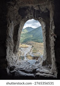View through a stone window showcasing a mountainous landscape with a river winding through greenery and a small settlement in the distance