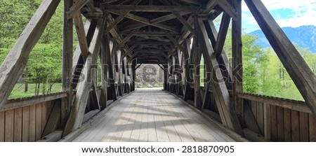View through the timber structure of a historic covered wooden bridge with repeating wooden beams, a plank walkway and strong central perspective. Green woodland and mountains are visible beyond the bridge entrance.