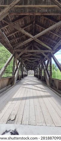 View through the timber structure of a historic covered wooden bridge with repeating wooden beams, a plank walkway and strong central perspective. Green woodland and mountains are visible beyond the bridge entrance.