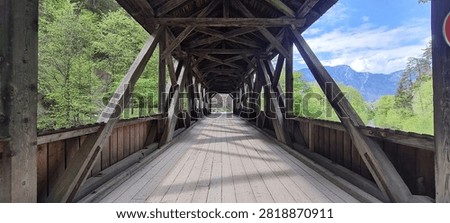 View through the timber structure of a historic covered wooden bridge with repeating wooden beams, a plank walkway and strong central perspective. Green woodland and mountains are visible beyond the bridge entrance.
