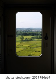 View through train window of fields, trees, and distant town under a clear sky.