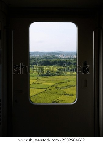 View through train window of fields, trees, and distant town under a clear sky.
