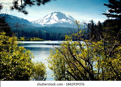 A view through trees across Manzanita Lake and its sparkling waters to a partly snow covered Lassen Peak within the Lassen Volcanic National Monument in Northern California.