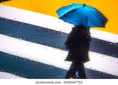 view through the window covered with raindrops on people under colored umbrellas crossing the pedestrian crossing, focus on raindrops on the glass